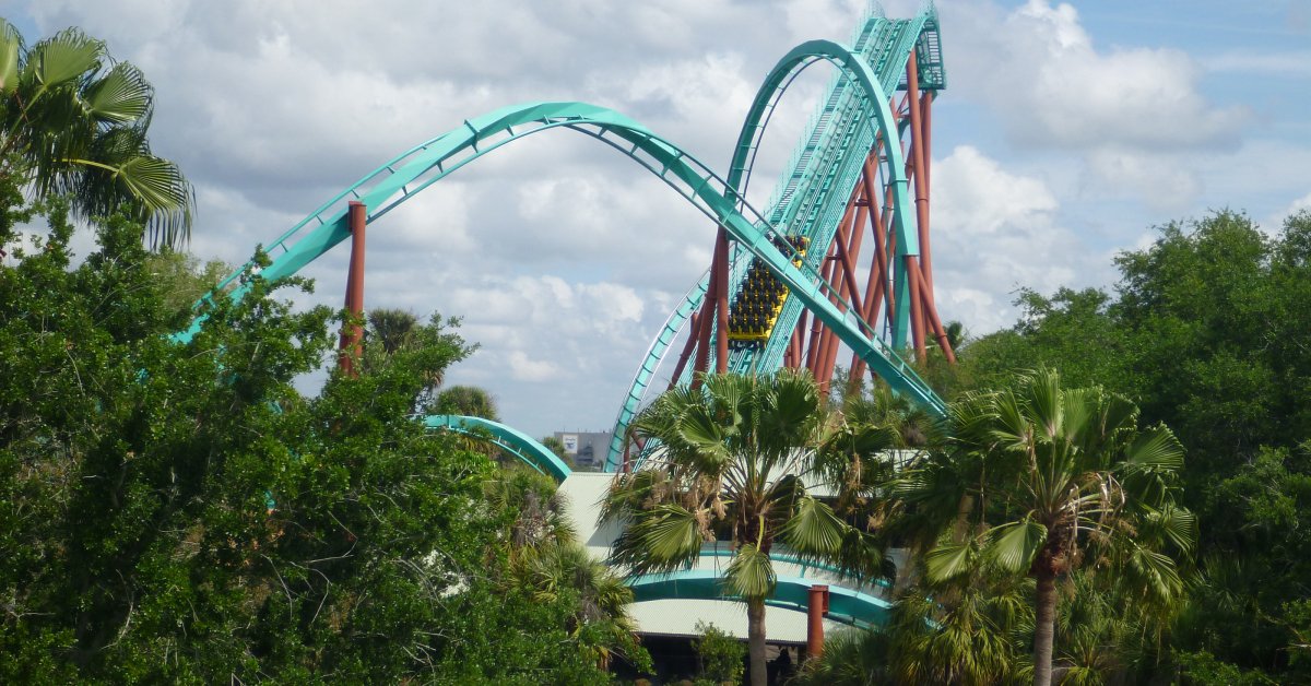 A cyan rollercoaster surrounded by tropical greenery, including palm trees. The skies are partly cloudy.
