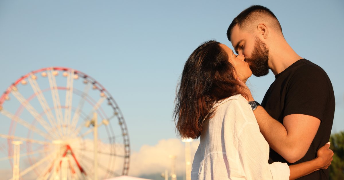A couple kissing at an amusement park at dusk. In the background are a large Ferris wheel and a white tent.