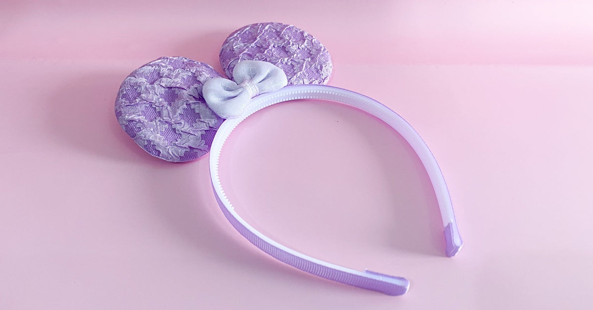A purple headband with a small white bow and large mouse ears. The headband is sitting against a pink background.