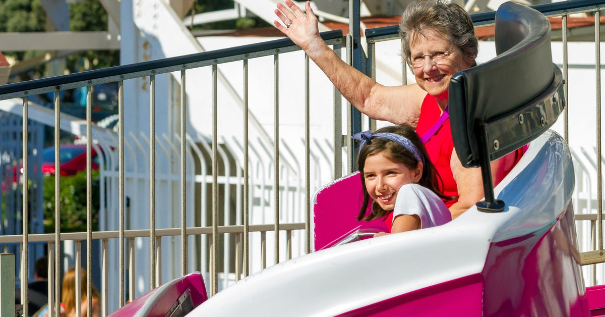 An elderly woman and a little girl riding on a theme park attraction. The elderly woman is waving, and the girl is smiling.