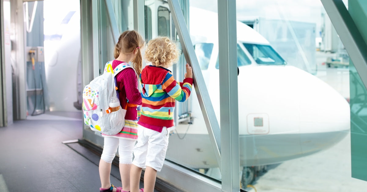 Two kids with luggage looking out the window at an airport. Outside, there is a large plane parked on the tarmac.