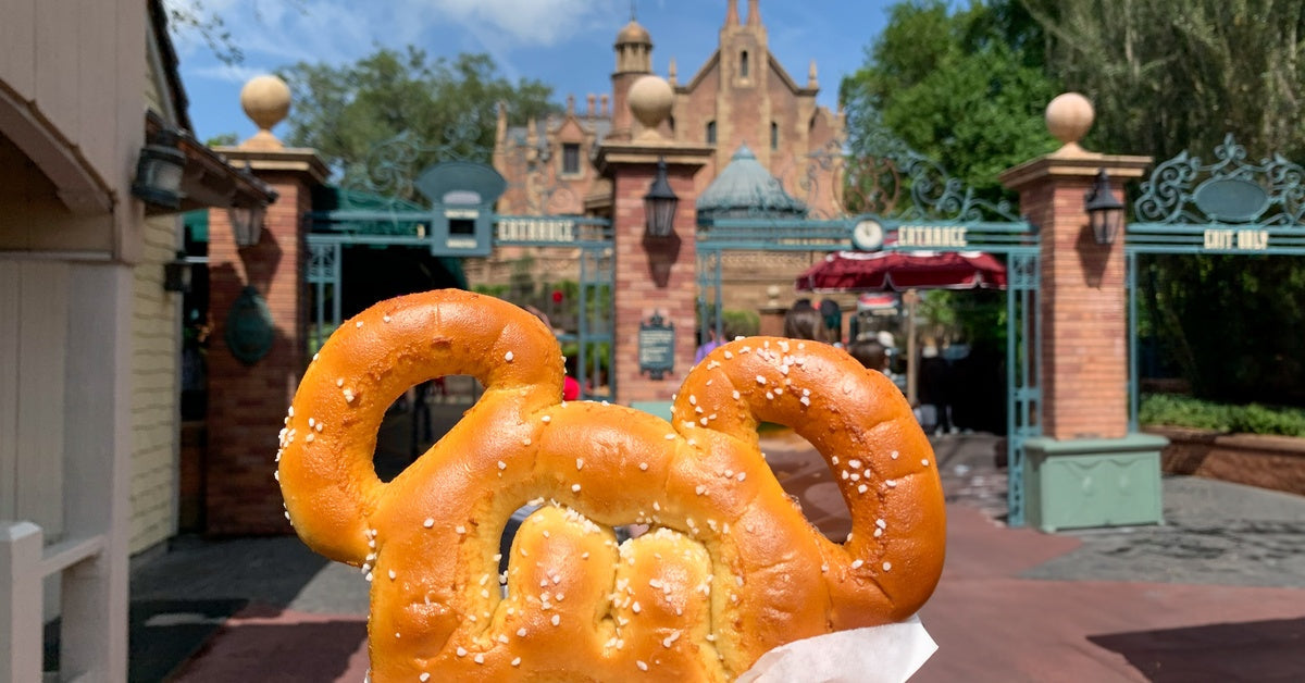 A person holding a pretzel in front of an amusement park entrance. The pretzel is in the shape of Mickey's head.