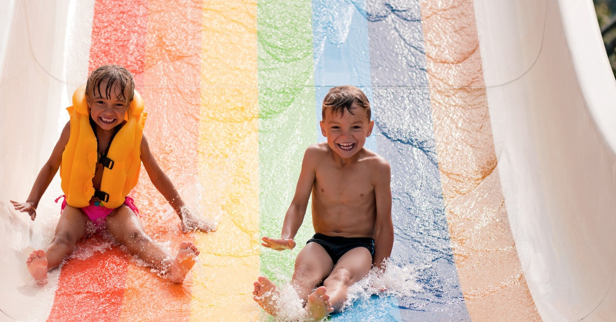 Two kids sliding down a water slide wearing swimsuits and a life jacket. There is a rainbow on the water slide.