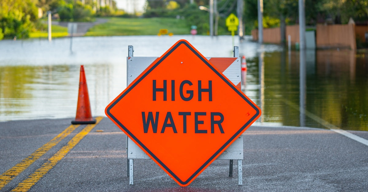 An orange construction sign that reads “HIGH WATER” in front of a flooded street. There is a cone behind the sign.