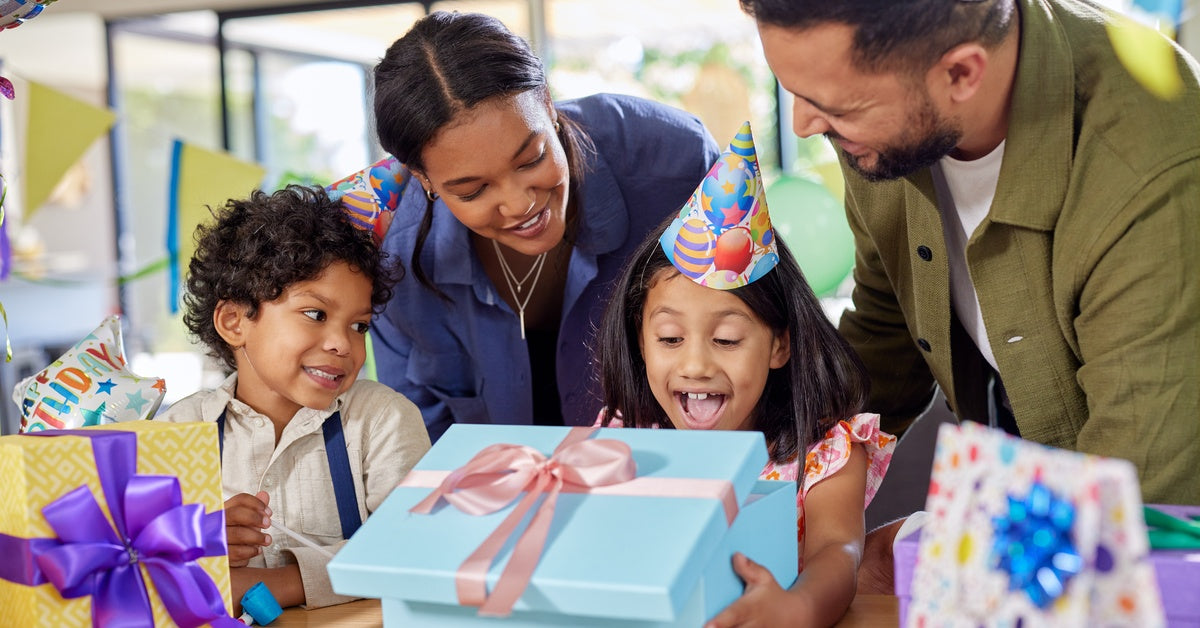 A family gathered around a young girl wearing a birthday hat. She opens a blue present in front of her.