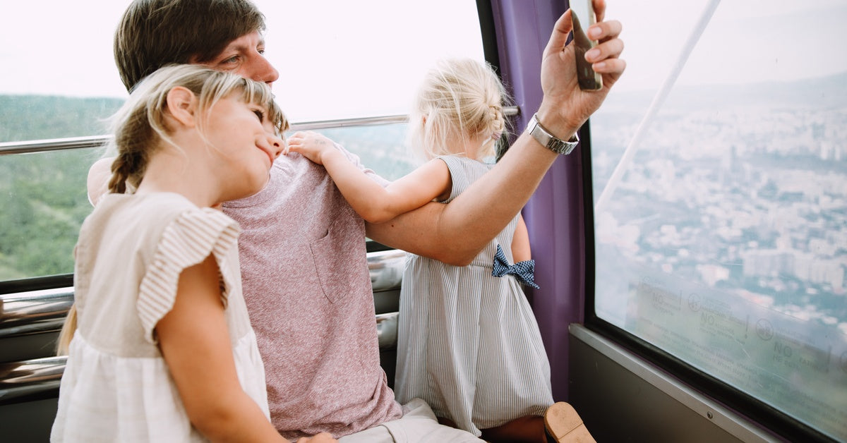A father and his two daughters riding on a Ferris Wheel attraction. The father is taking a photo of his family.