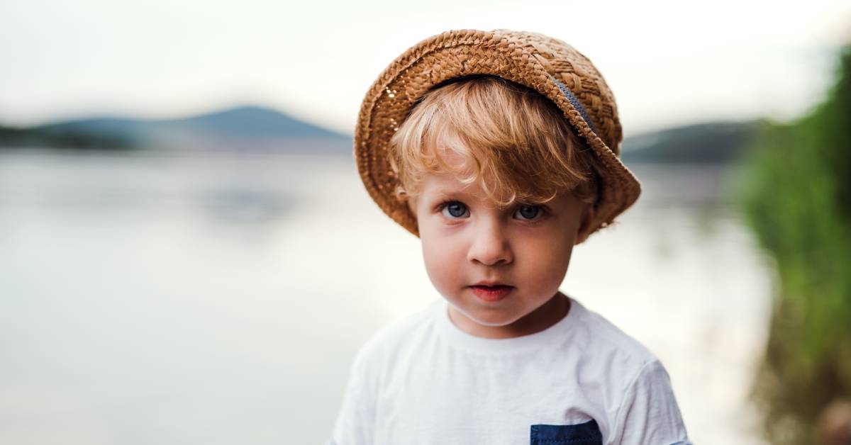 A child stands alone outside wearing a hat and a white T-shirt and there is a tall green bush nearby.