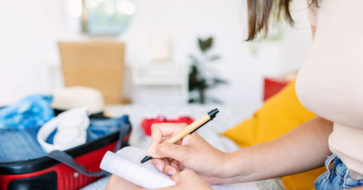 A woman making a travel checklist in a notebook. She is marking and checking her items with her luggage open.