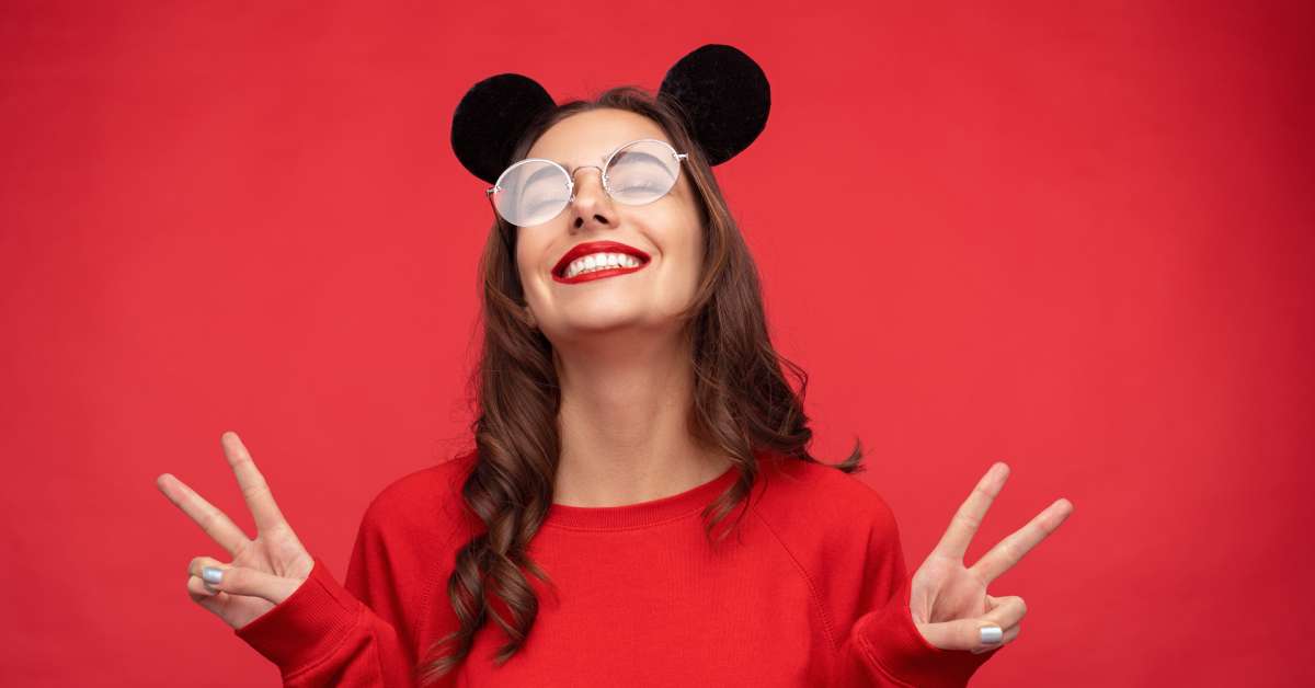 A girl wearing a headband with black mouse ears, posing with two peace signs. She is also wearing a red sweater.