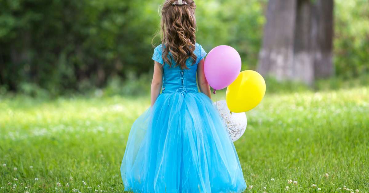 A little girl holding balloons in a princess gown. The dress is a light blue with frilly tulle as the skirt.