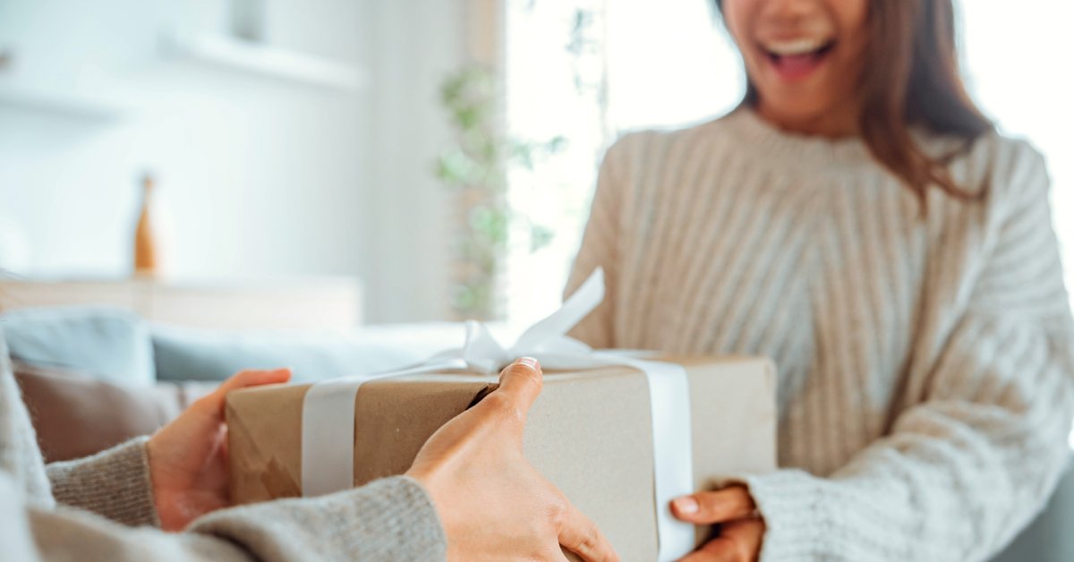 A woman receiving a wrapped present from another person. She is excited as she grabs the box with both hands.