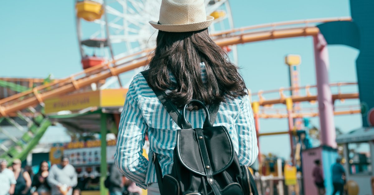 A woman with her back turned away facing a theme park. She is wearing a straw hat and a black backpack.