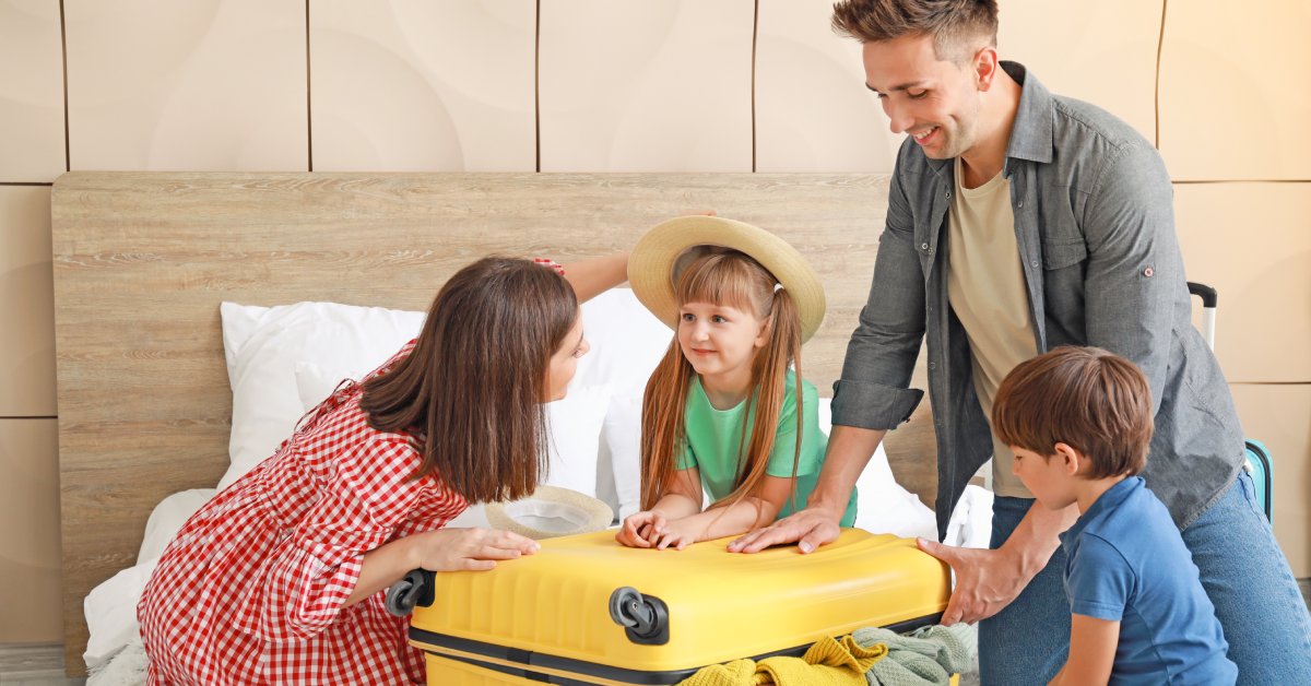 A family of four packing a yellow suitcase on top of a bed. The suitcase is overflowing with clothes as they push it down.