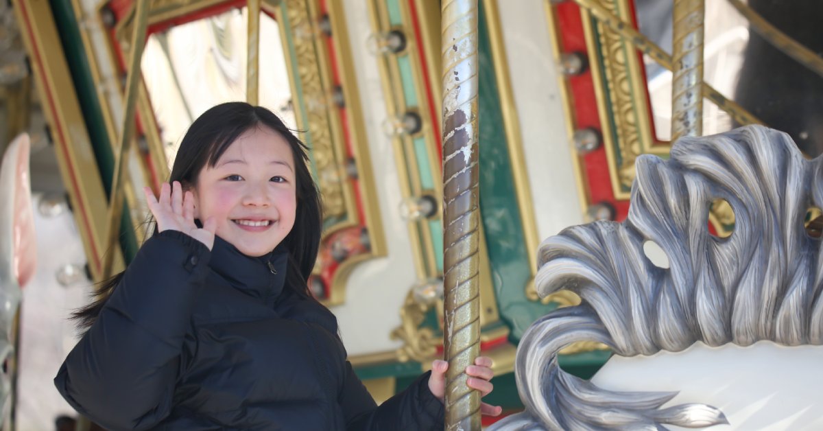 A little girl riding on a carousel. She is wearing winter clothes with one hand on the ride and the other waving.