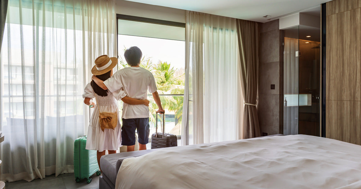 A couple inside a hotel room holding their rolling luggage with one hand. They look outside their hotel window.