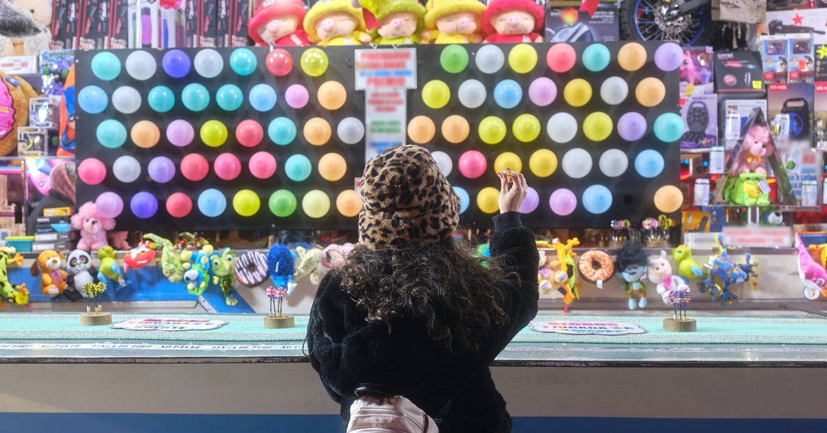 A woman holding a dart in front of a carnival game. There is a wall of balloons with many stuffed animals as prizes.