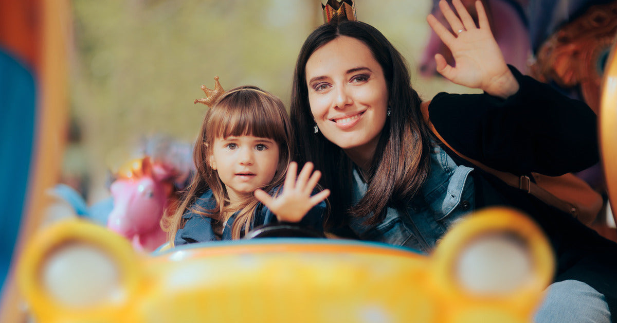 A mom and her daughter waving while riding on a park attraction. They are sitting in a yellow cart on the ride.