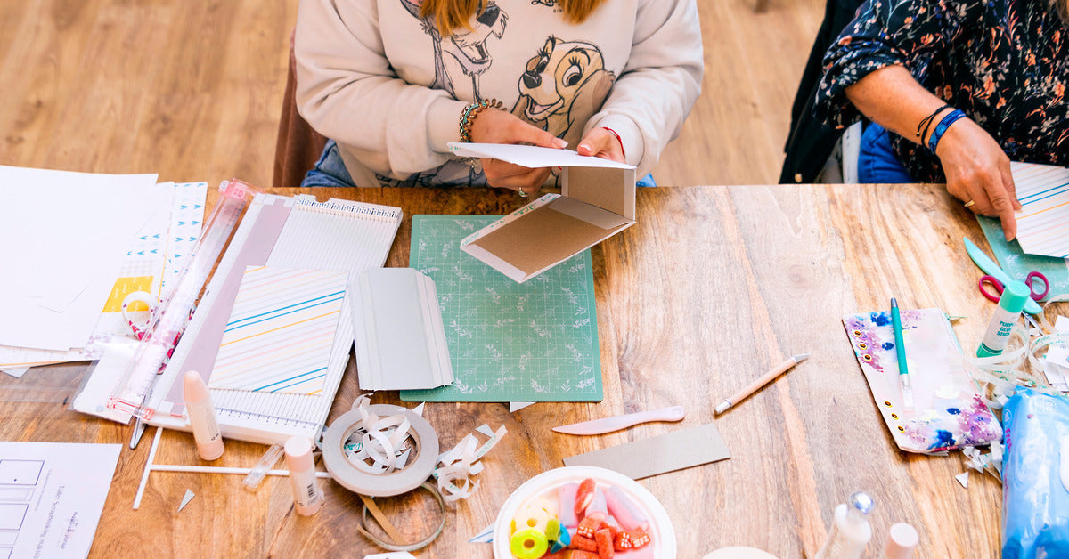 Two people sitting at a wooden table with craft supplies scattered across the table. They are making a scrapbook.