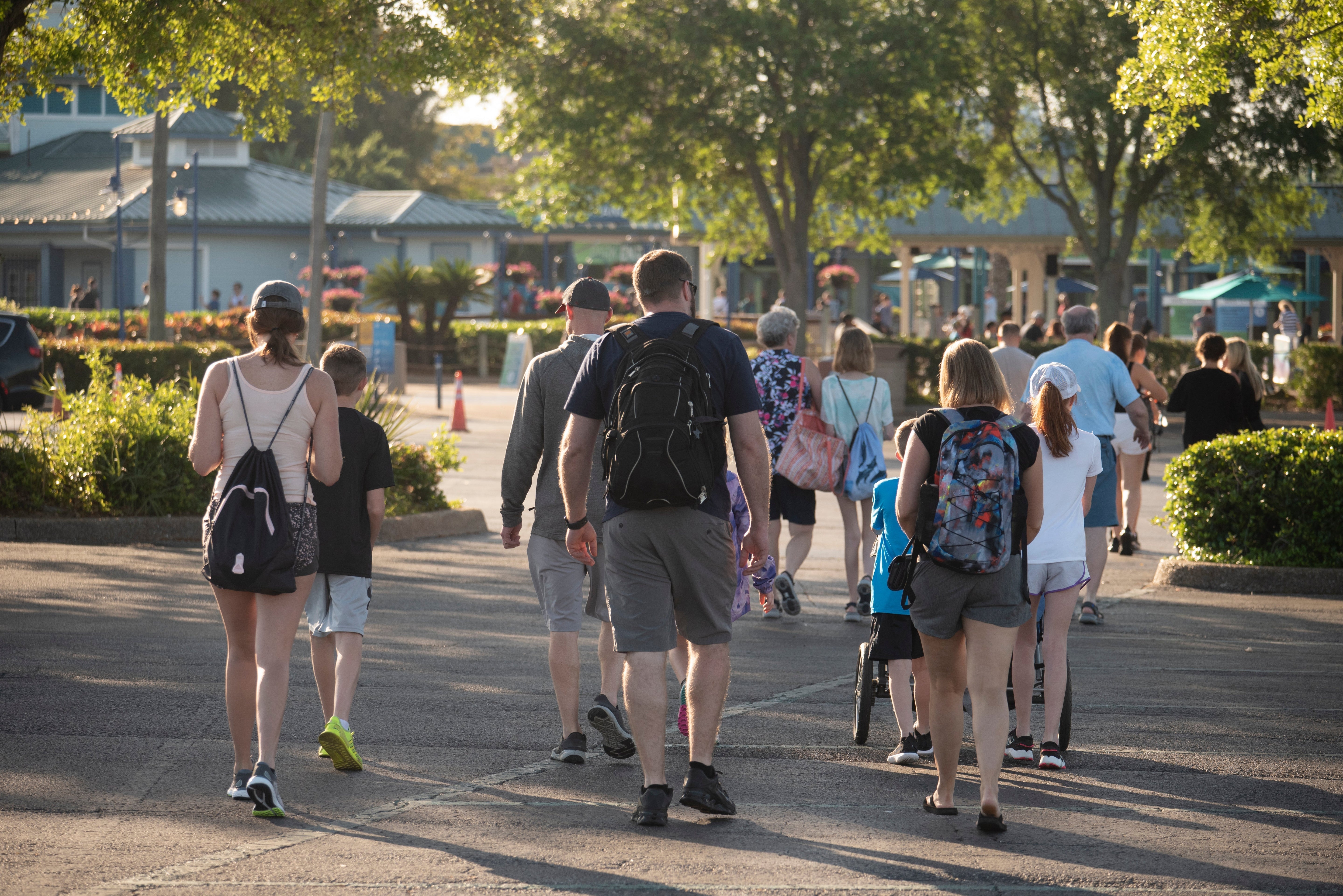 A group of people walking toward the front entrance of a theme park. Many of them are wearing backpacks and hats.