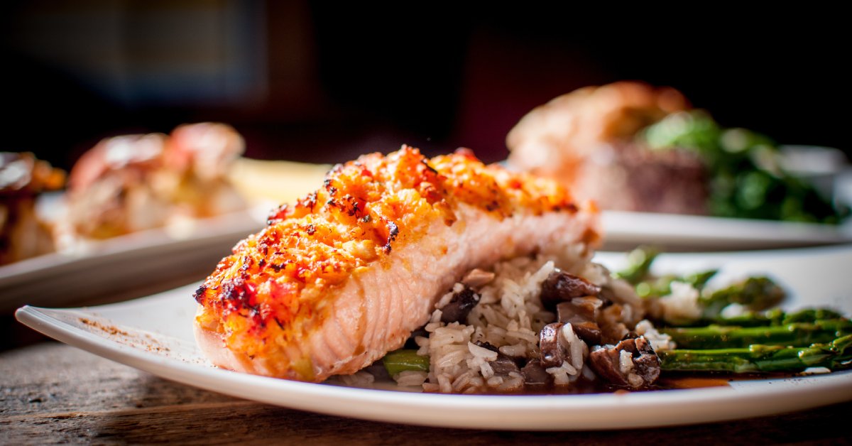 Crusted salmon with rice pilaf and grilled asparagus served on a white plate. There are other dishes in the background.