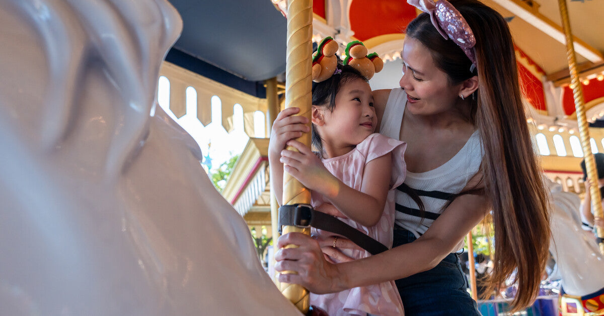 A mother and her daughter sitting on a fake horse, riding a carousel. They wear stylish mouse ear headbands.