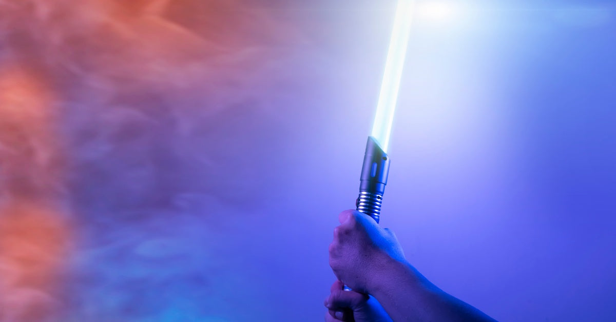 Two hands holding an illuminated blue light saber by the handle. There is blue and red smoke in the background.