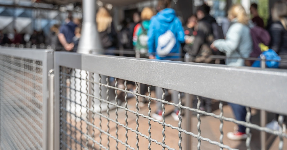 People standing in a queue line divided by a metal fence. Many of them are wearing backpacks as they’re waiting in line.