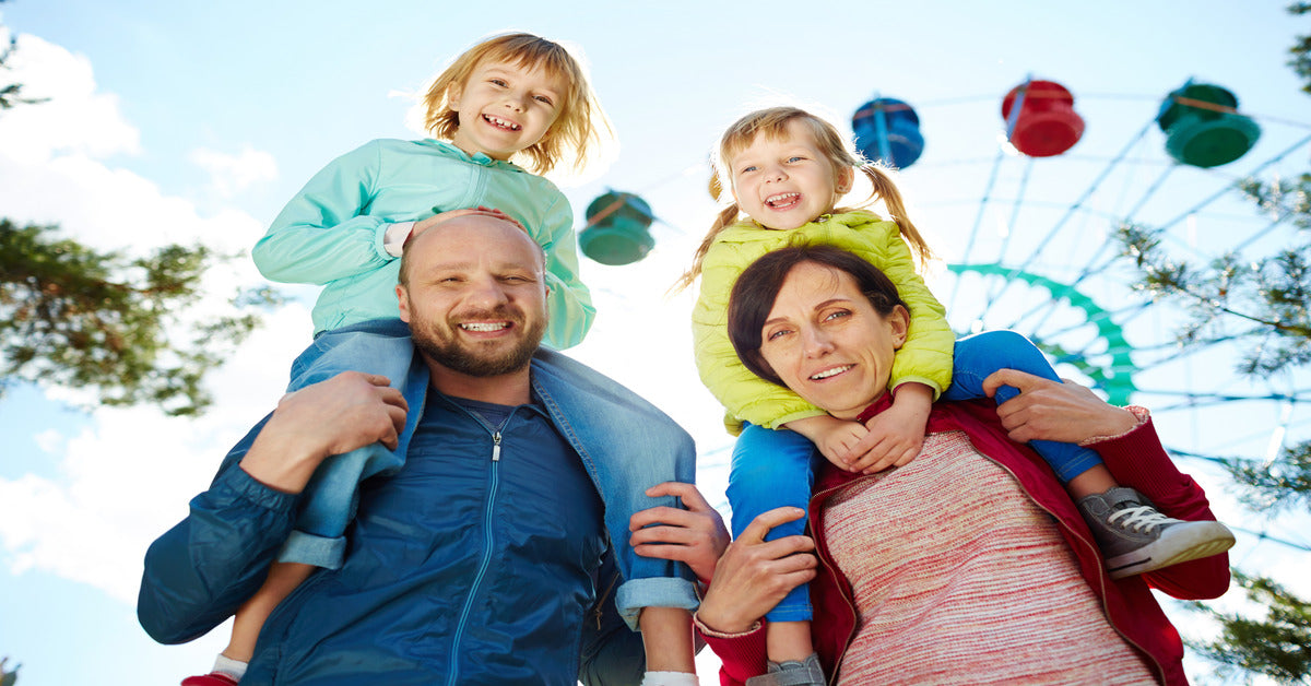 A mother and father carrying their children on their shoulders. There is a colorful Ferris wheel behind them.