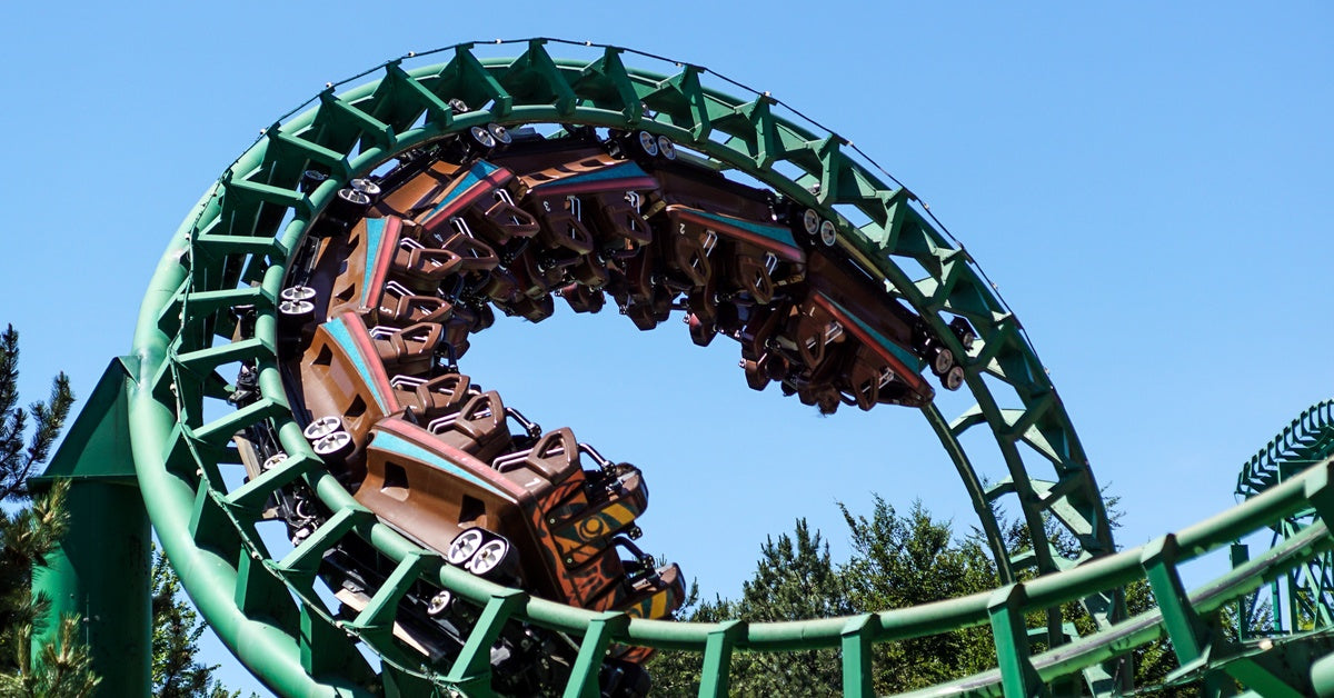 A green, metallic roller coaster with brown cars on the track. There are people riding in the seats, driving through a loop.