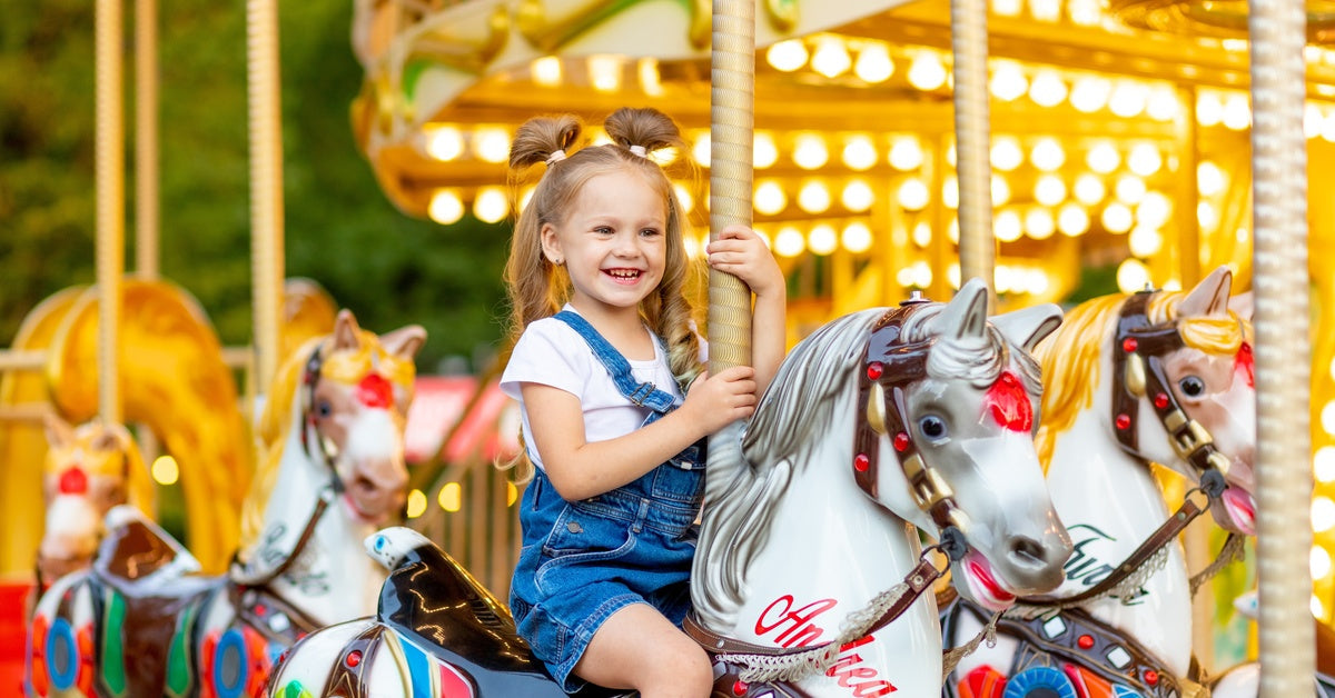 A little girl wearing denim overalls riding on a carousel at an amusement park. She sits on a white horse.
