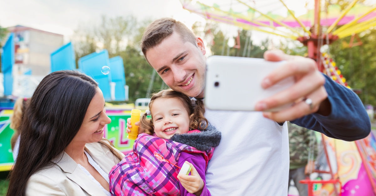 A family using their cell phone to take a photo at a theme park. The parents hold their daughter in their arms.