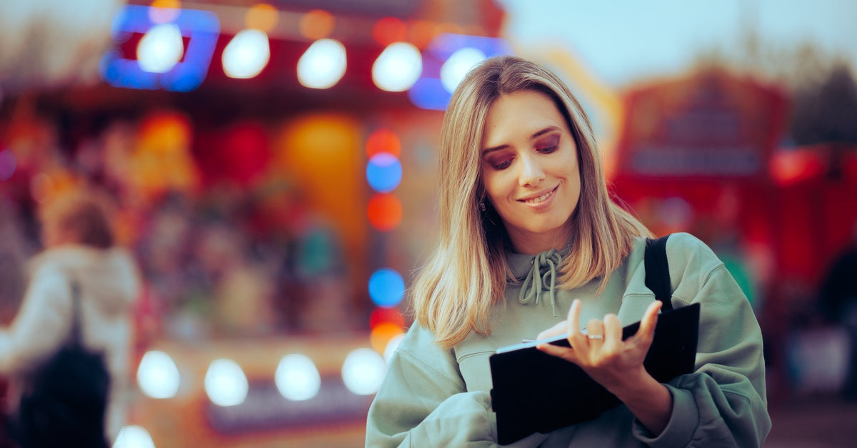 A woman looking inside a black notebook at an amusement park. She is smiling as she reads the page in front of an attraction.