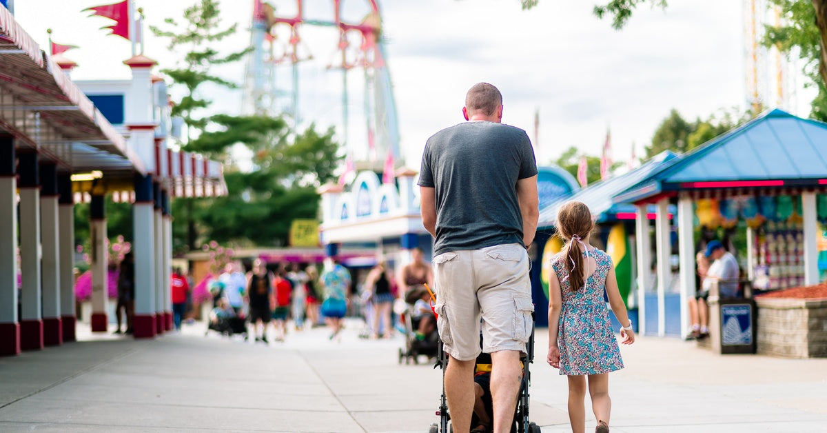A father and his daughter walking through an amusement park. The father is pushing a baby stroller in front of him.