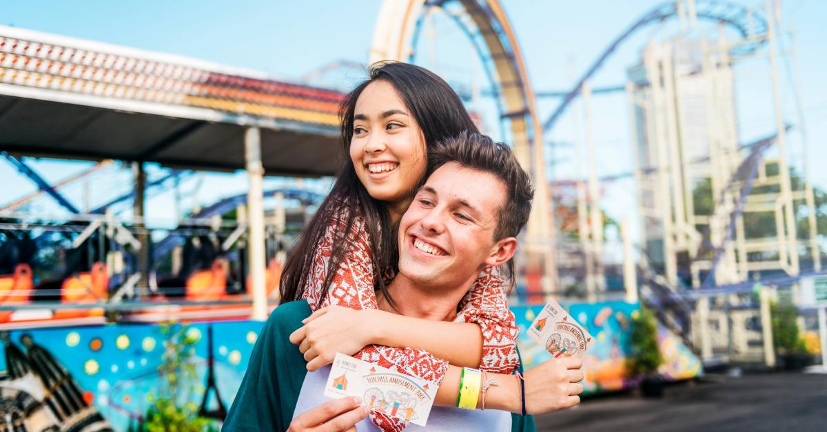 A man carrying a woman on his back at a theme park. They are both holding admission tickets in front of them.
