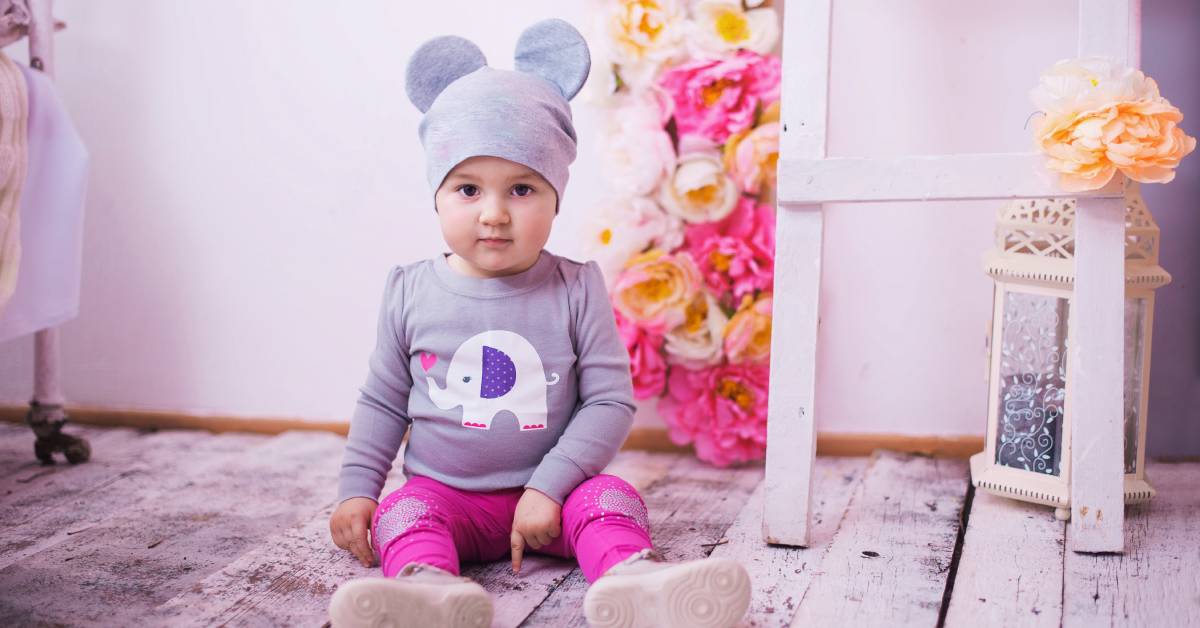 A young toddler wearing a gray hat with mouse ears, sitting on a wooden floor in front of colorful flowers and other decor.