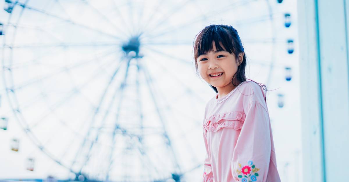 A young toddler smiling and wearing a pink outfit at a theme park while a Ferris wheel is visible behind them.