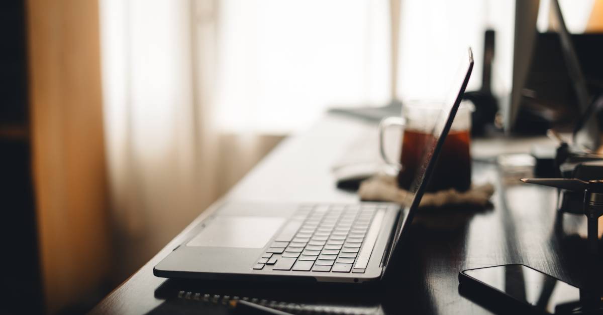 A dark desk in a home office sitting by a window while holding a laptop, a pitcher of tea, and random desk accessories.